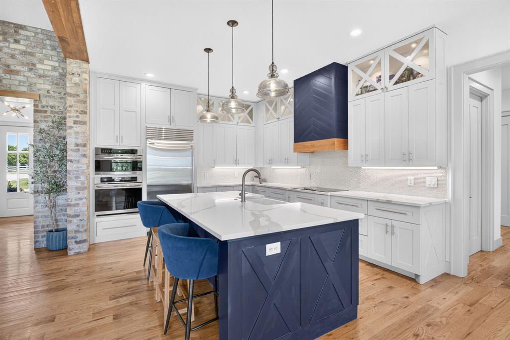 3008 Perkins Weatherford, TX 76088 - Photo 12 of 36 Gourmet kitchen featuring a large central island with a quartz countertop and navy blue base cabinetry, light wood-finish flooring, white perimeter cabinetry, stainless steel built-in appliances, and a unique range hood with a chevron pattern