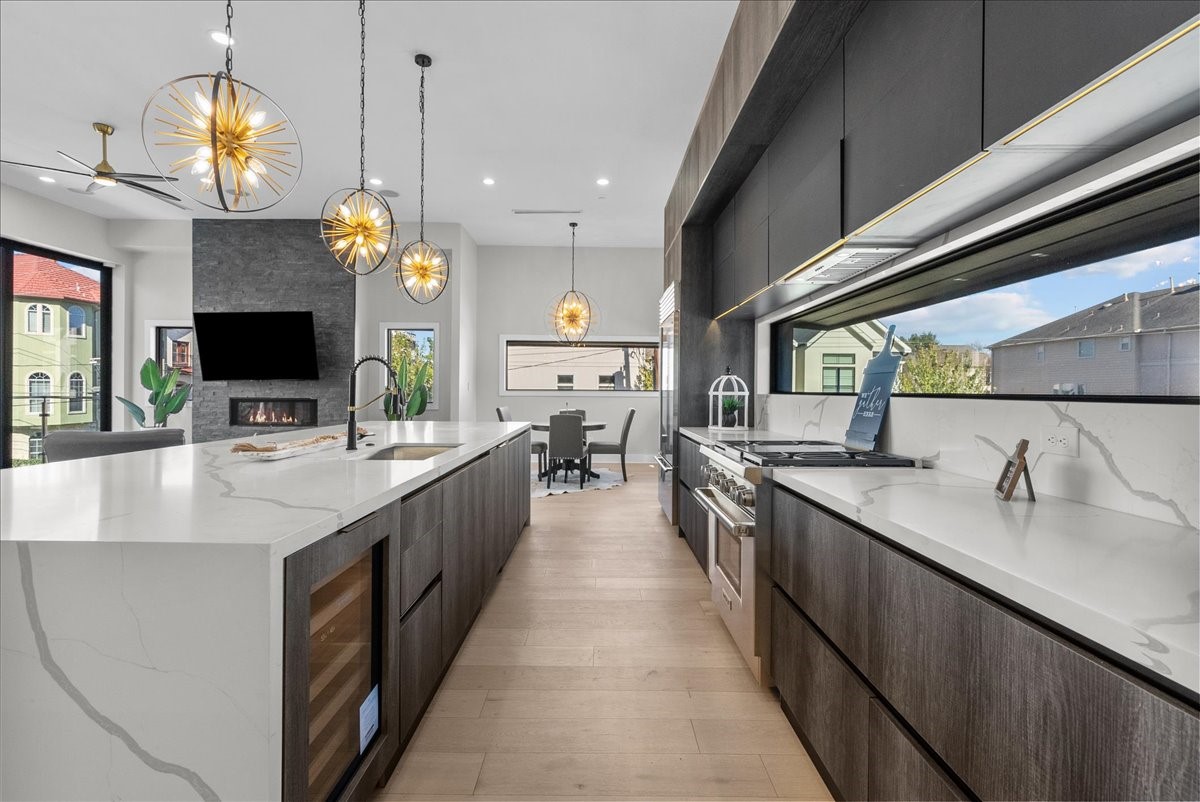 516 Reinerman Street Houston, TX 77007 - Photo 12 of 42 This modern kitchen features sleek dark cabinetry, marble countertops, and elegant pendant lighting. It includes a spacious island with a sink, a built-in wine cooler, and large windows providing abundant natural light. The open layout flows into a dining area, complemented by a cozy fireplace and contemporary decor.