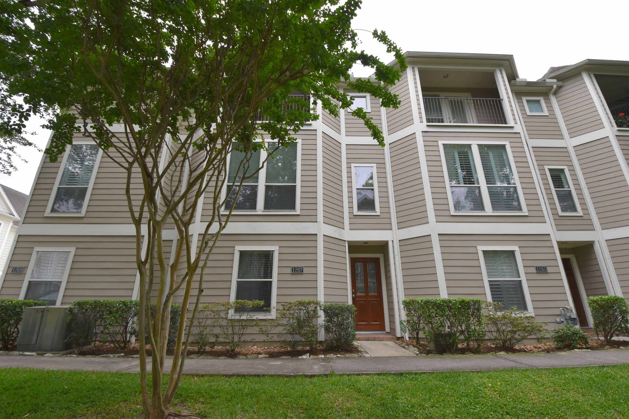 1267 West 17th Street Houston, TX 77008 - Photo 1 of 22 This is a multi-story townhouse with a beige exterior, featuring multiple windows, a central entry door, and is surrounded by green landscaping including a mature tree in the front yard.