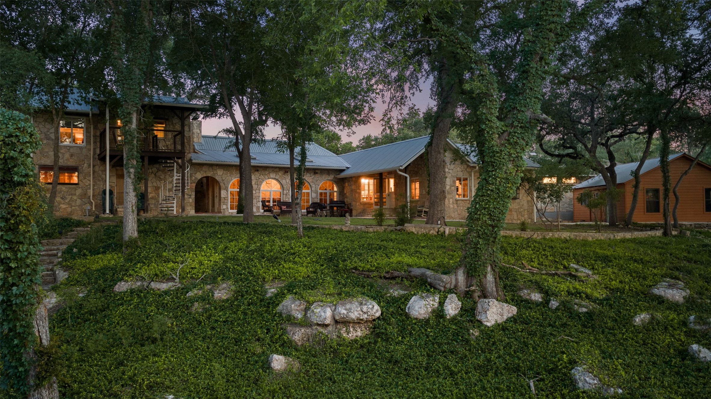 505 Red Corral Ranch Road Wimberley, TX 78676 - Photo 24 of 40 A grand stone dwelling with arched windows and a spiral staircase, nestled amongst mature trees and lush greenery.