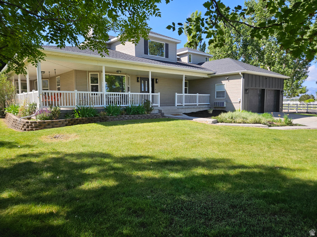 812 East Bates Cyn Road Tooele, UT 84074 - Photo 137 of 144 Two-story residence featuring a full-width covered front porch with white railings, multiple dormer windows, an attached two-car garage with black doors, and a stone-block retaining wall with landscaping