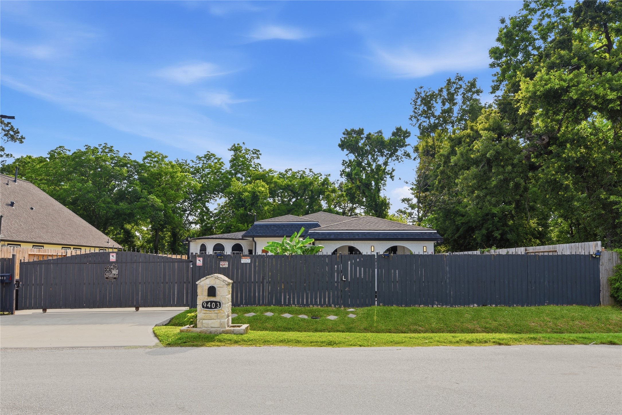 9403 Spaulding Street Houston, TX 77016 - Photo 1 of 50 This photo depicts a single-story home with a dark fence and a gated driveway. The house is surrounded by mature trees, offering privacy and a natural setting. The front yard is neatly maintained, and there’s a stone mailbox with the house number.