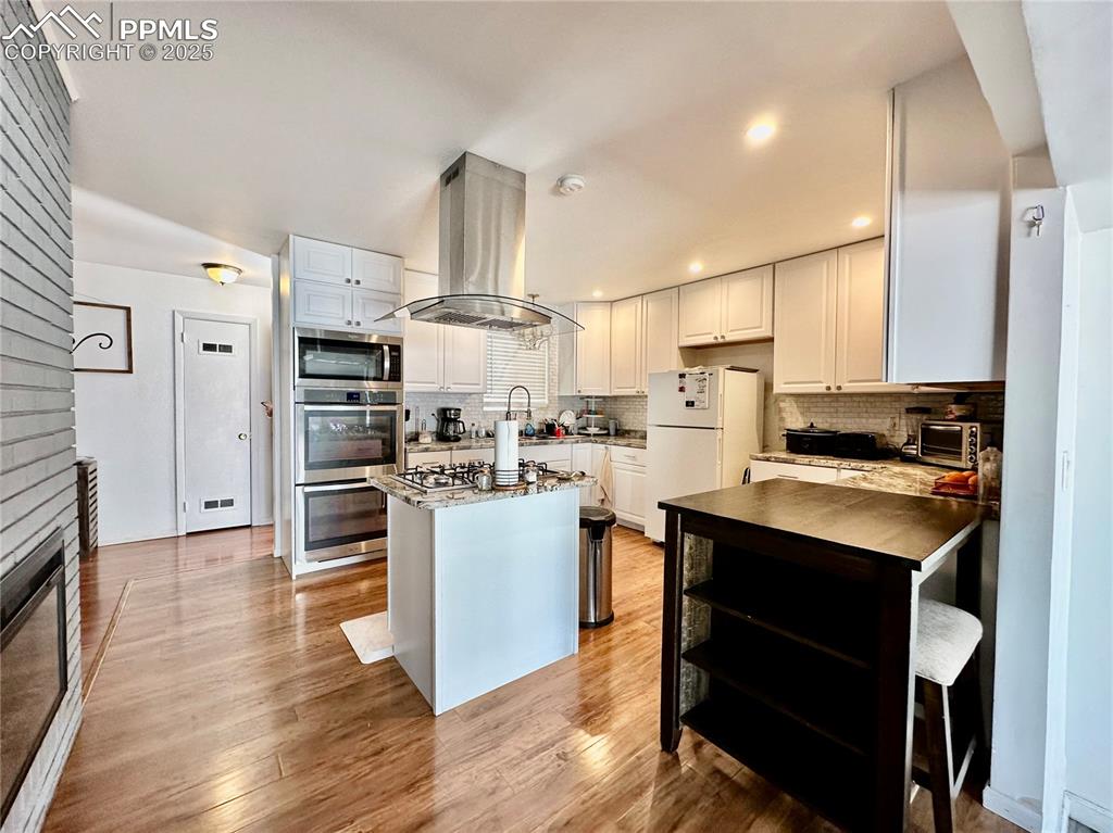 1701 Jerry Murphy Road Pueblo, CO 81001 - Photo 7 of 20 Kitchen featuring island range hood, stainless steel appliances, backsplash, white cabinets, and light wood-type flooring