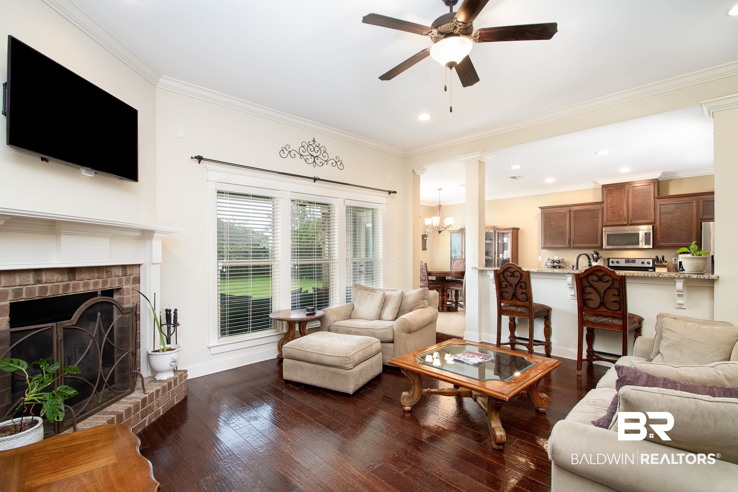 8625 Rosedown Lane Daphne, AL 36526 - Photo 7 of 29 Living room featuring a brick fireplace, ceiling fan with notable chandelier, crown molding, and dark wood-type flooring