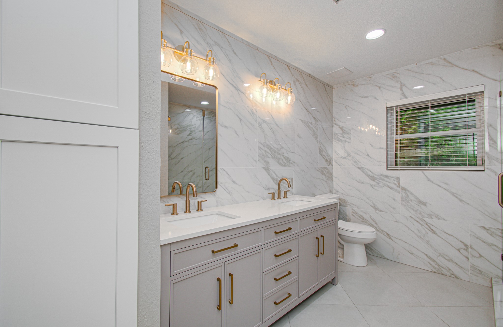 1801 Benbow Way Houston, TX 77080 - Photo 22 of 32 This bathroom features a sleek, modern design with marble-patterned walls and floors. It includes a double vanity with gold fixtures, elegant lighting, and a large mirror, creating a luxurious and bright atmosphere.