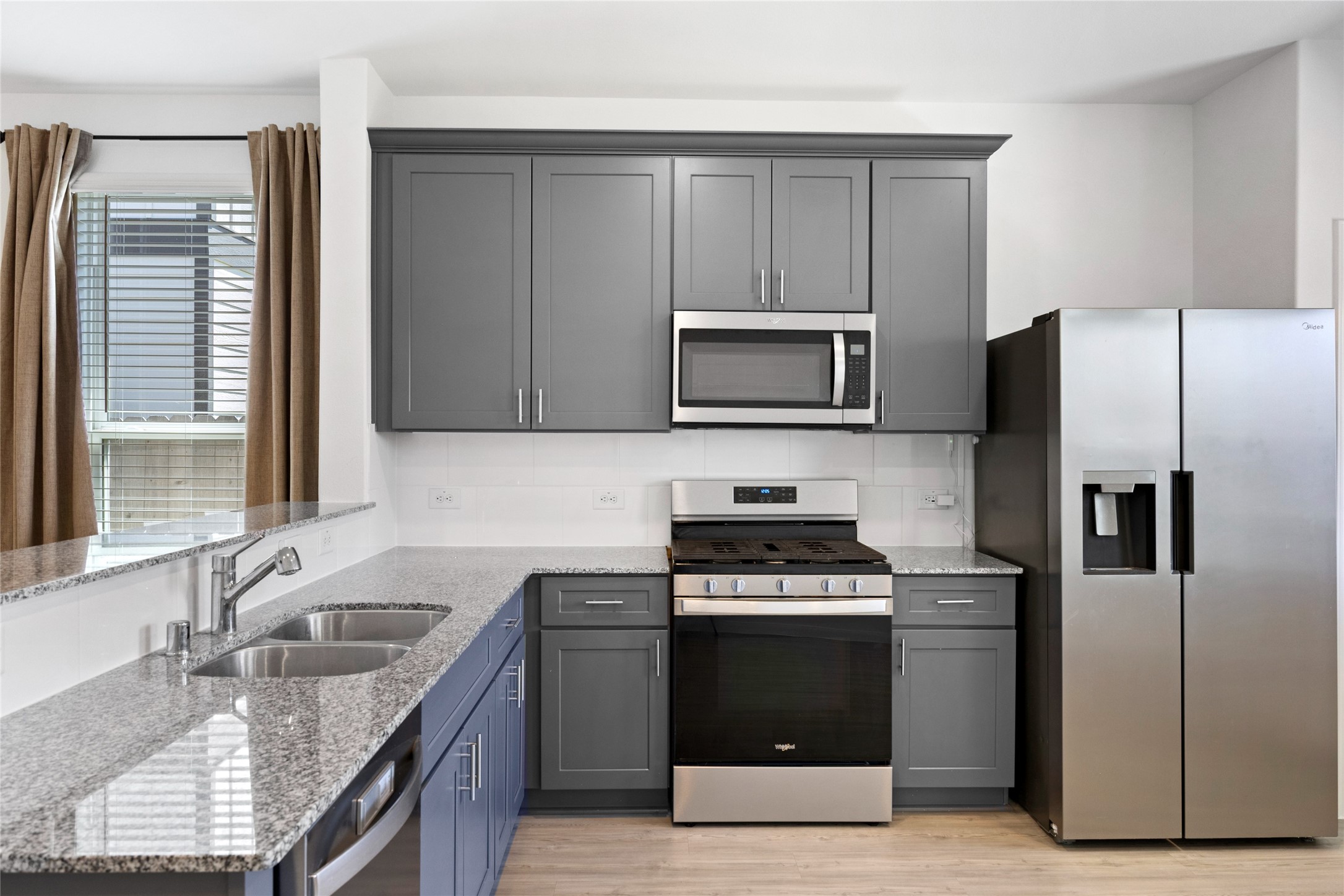 17907 Grain Auger Road Hockley, TX 77447 - Photo 14 of 24 modern kitchen features sleek gray cabinetry, stainless steel appliances, a granite countertop, and a double sink. The space is brightened by a window with curtains, offering a stylish and functional cooking area.