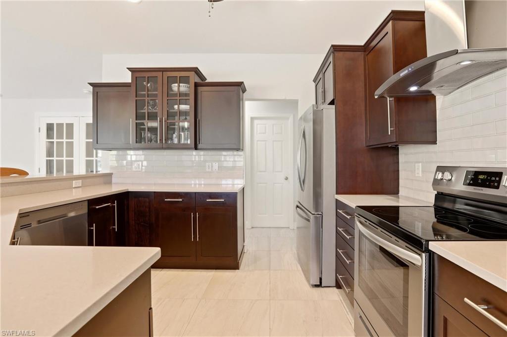 4282 Longshore Way North Naples, FL 34119 - Photo 10 of 43 Kitchen with stainless steel appliances, glass fronted cabinets, dark wood finish cabinetry, backsplash, and light stone countertops