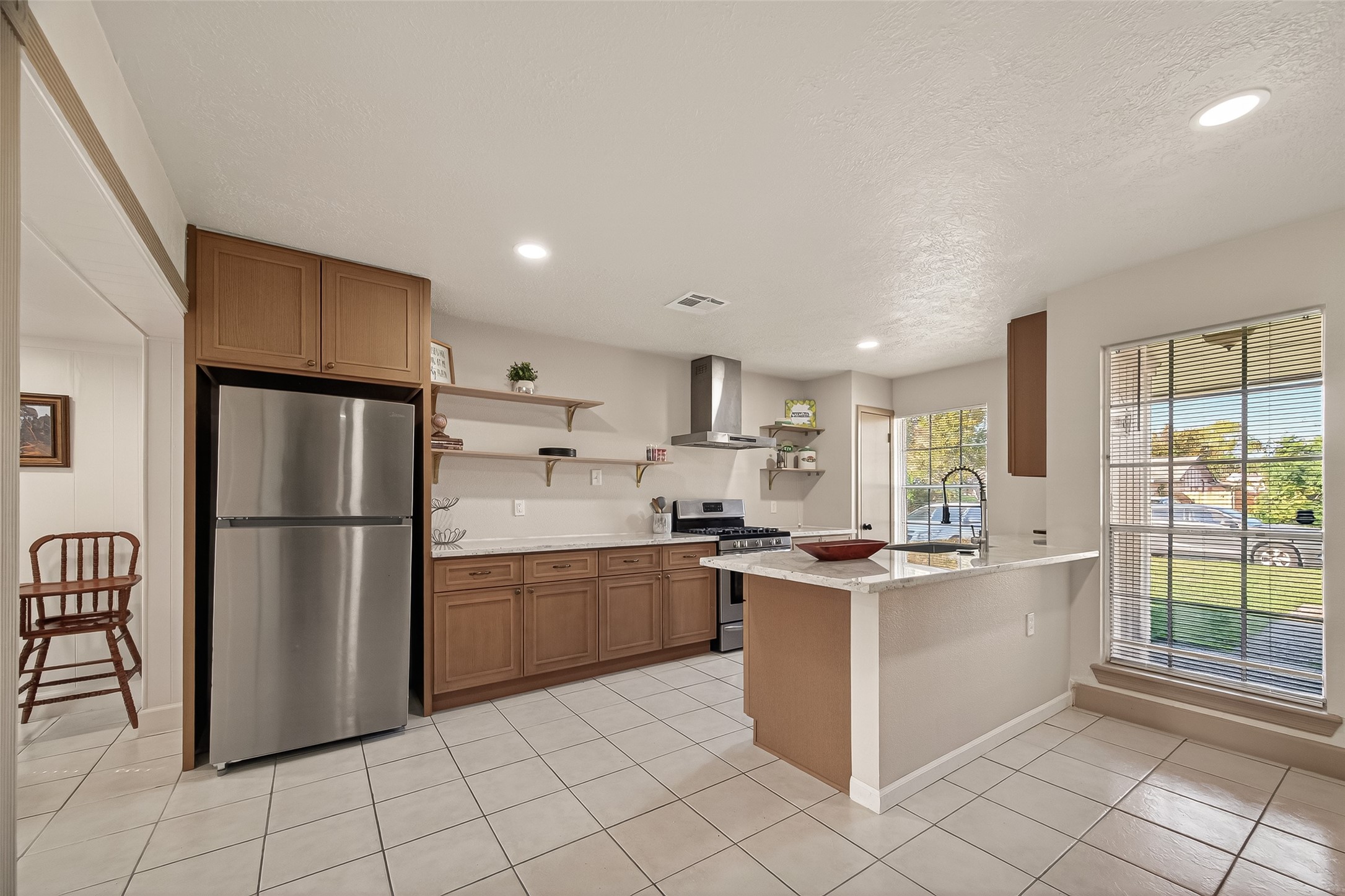 This kitchen features modern stainless steel appliances, ample cabinetry, and an island with a breakfast bar. Large windows let in plenty of natural light, and the open layout connects to an adjacent dining area, creating a welcoming and functional space.