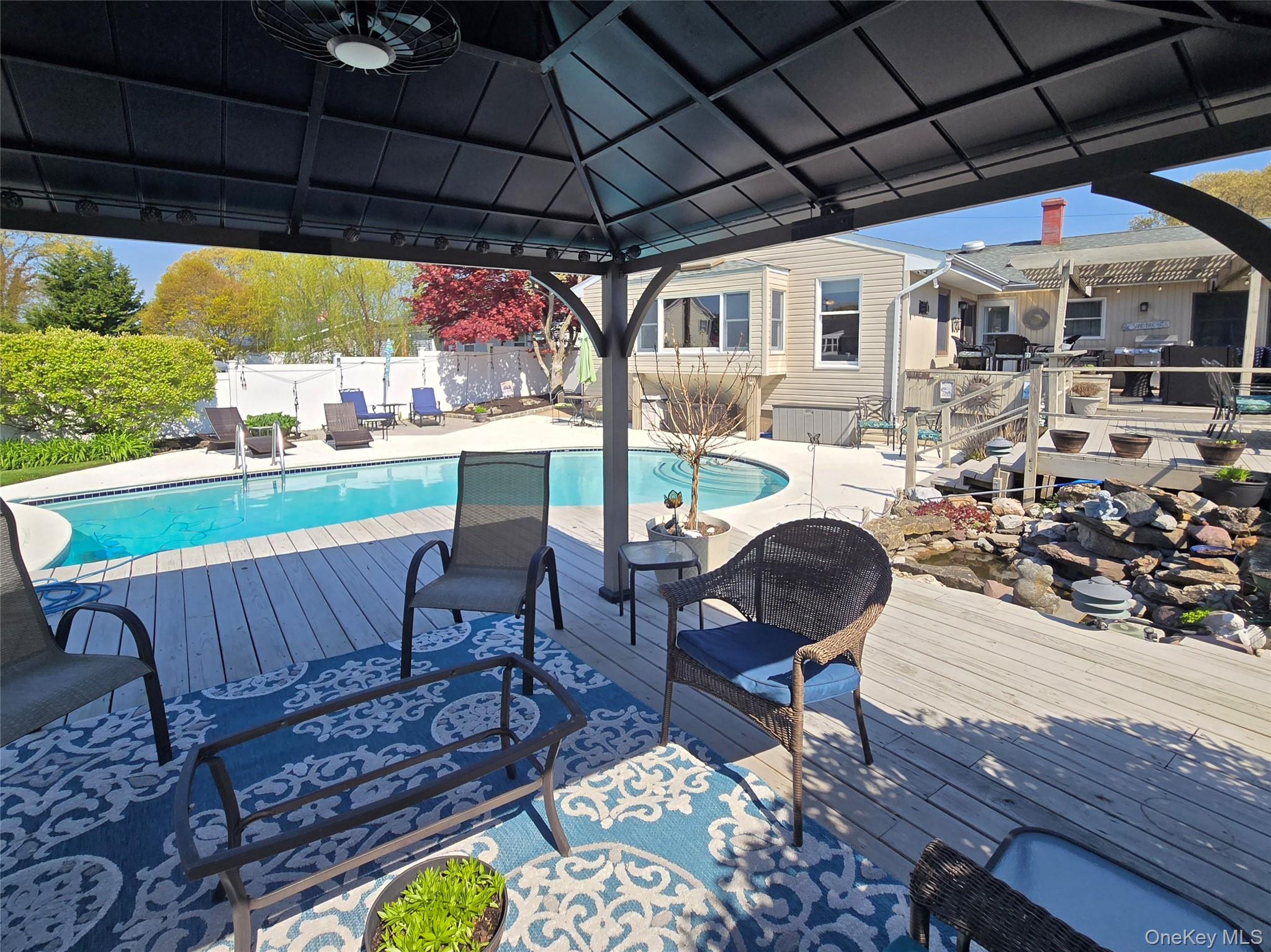 212 Matthews Road Oakdale, NY 11769 - Photo 33 of 38 Covered outdoor living space featuring a ceiling fan, composite decking, a swimming pool, a rock garden, and a white privacy fence