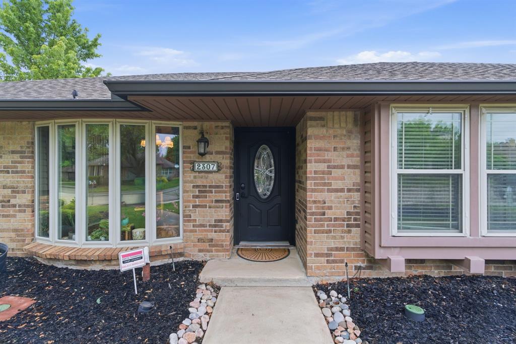 2307 Castle Rock Road Carrollton, TX 75007 - Photo 4 of 34 The property features a brick exterior with a dark front door and decorative glass insert, complemented by a bay window and additional windows with blinds