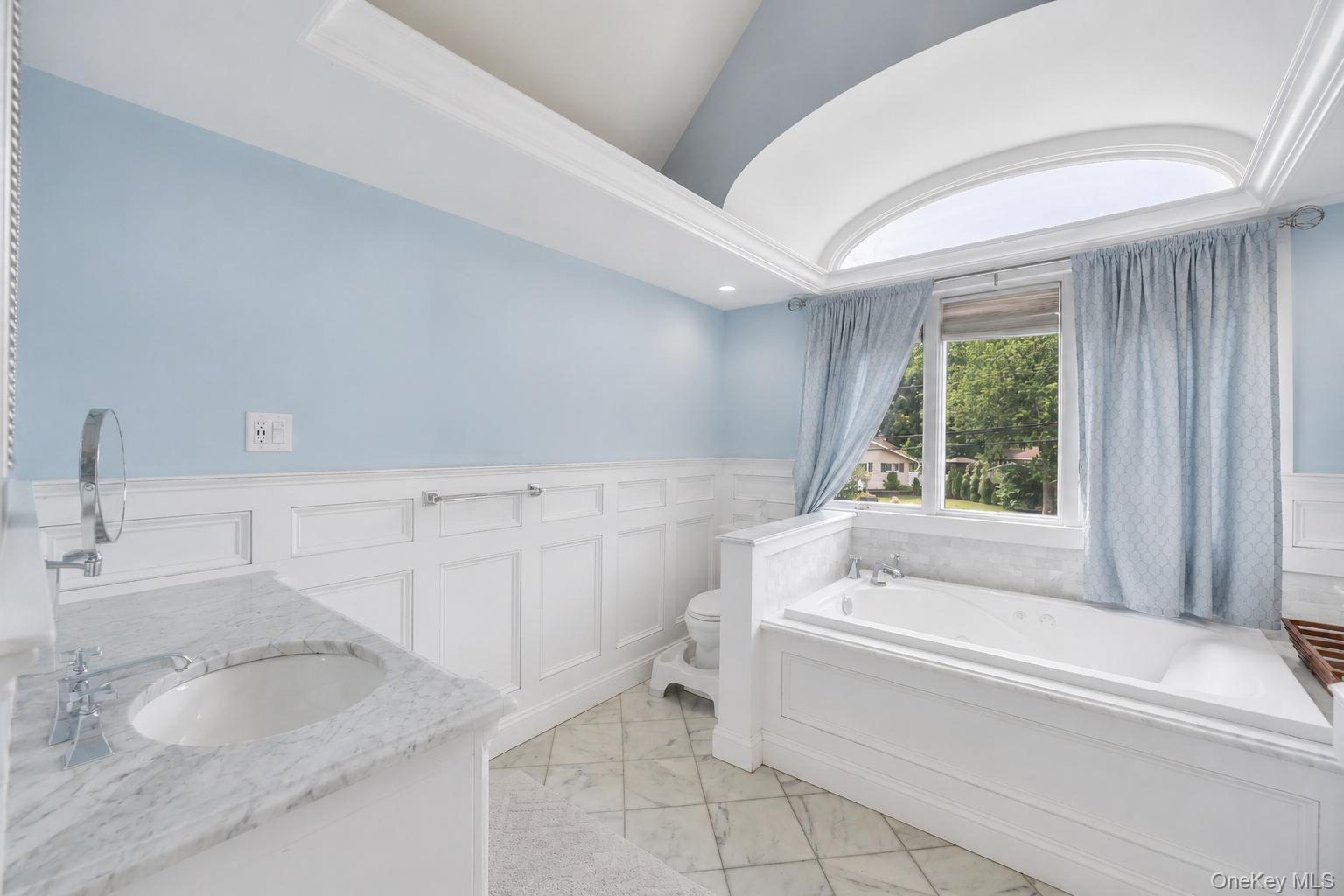 19 Joan Road Bay Shore, NY 11706 - Photo 27 of 50 Bathroom featuring a spacious built-in soaking tub, an arched transom window, a single vanity with a stone countertop and undermount sink, wainscoting, and large format floor tiles
