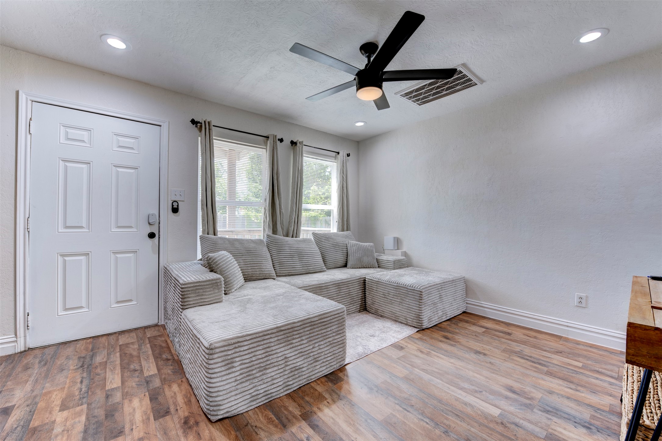 1121 Panama Street Houston, TX 77009 - Photo 5 of 35 Living room wide angle showcasing white six-panel entry door with smart lock, oversized window with grommet linen curtains, wood-look LVP flooring, five-blade black ceiling fan with light kit, recessed lighting, and oversized returns with white baseboard trim throughout.