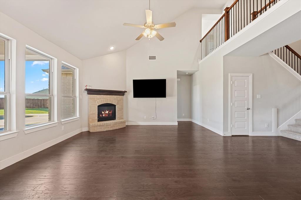Vaulted ceiling with a ceiling fan, corner fireplace with a brick surround, multiple windows, wood-finish flooring, and a second-story interior balcony with wood and metal railing