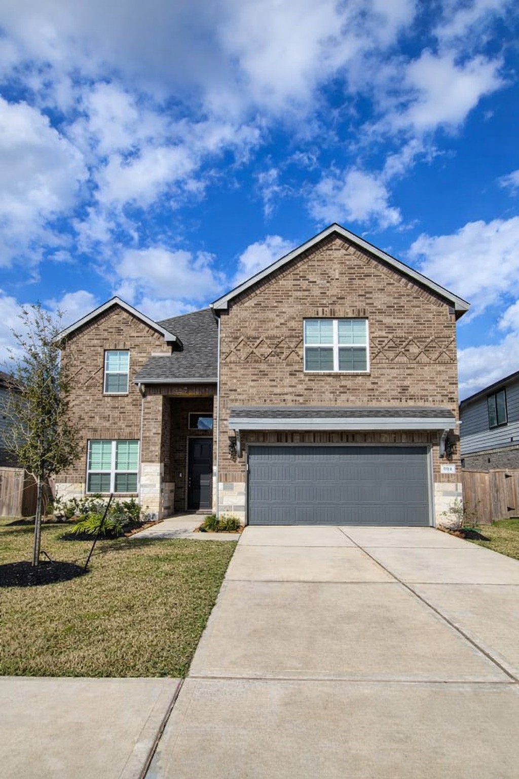 This photo showcases a modern two-story brick house with a gabled roof and a two-car garage. The driveway is spacious, and the front yard is neatly landscaped with a small tree and plants. The house features large windows, providing plenty of natural light.