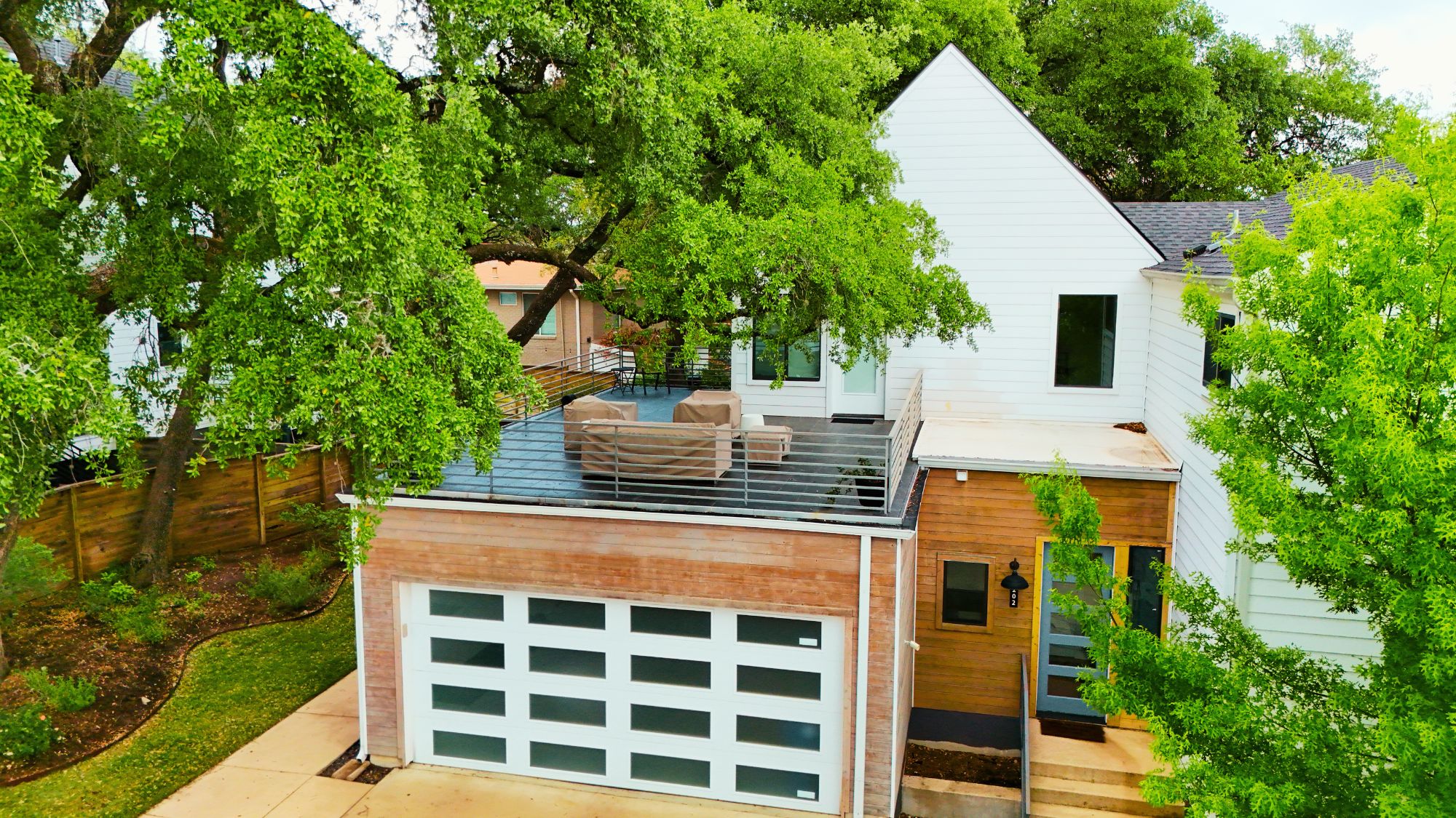 Contemporary exterior featuring white siding, natural wood accents, a two-car garage with modern glass-paneled doors, a private rooftop deck with metal railings, and a prominent front entry with a glass-paneled door