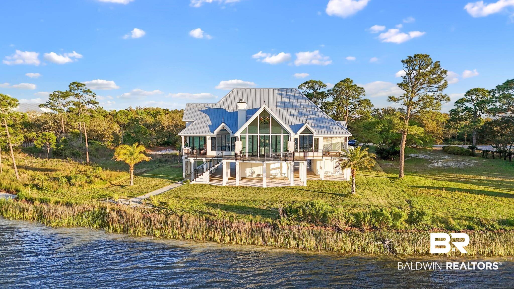 33349 River Road Orange Beach, AL 36561 - Photo 43 of 61 Waterfront residence featuring a striking blue metal roof, an elevated foundation, expansive glass windows, multiple outdoor balconies, and surrounding mature trees