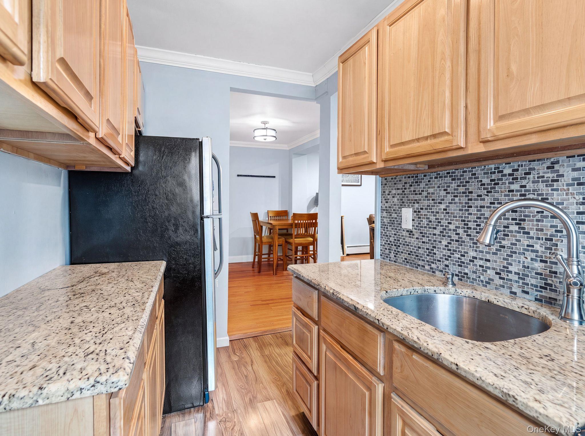 755 Bronx River Road, Unit 4B Bronxville, NY 10708 - Photo 14 of 27 Compact kitchen featuring wood-finish cabinetry, granite-style countertops, a mosaic tile backsplash, and a window providing natural light