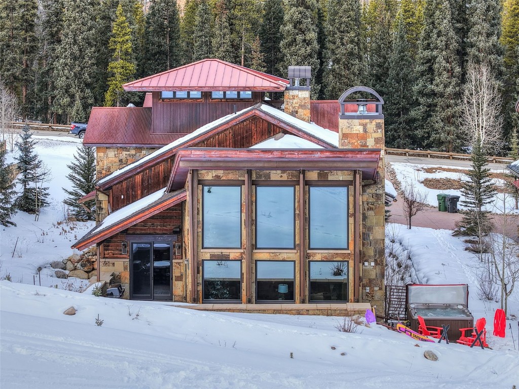 Snow covered back of property with a chimney, stone siding, and a wooded view