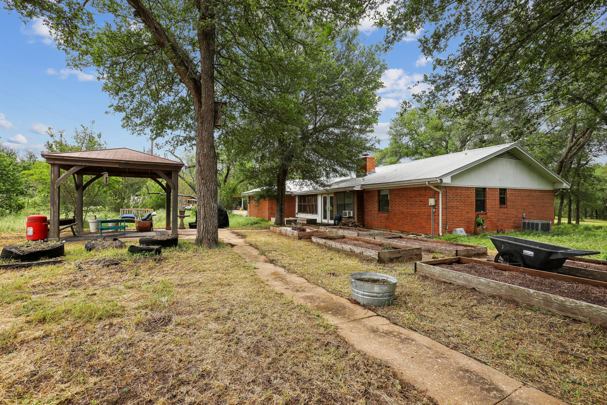 406 Monkey Road Elgin, TX 78621 - Photo 26 of 39 Brick exterior residence with a white gable roof, featuring a covered outdoor gazebo with a metal roof, multiple raised garden beds, and mature trees providing natural shading