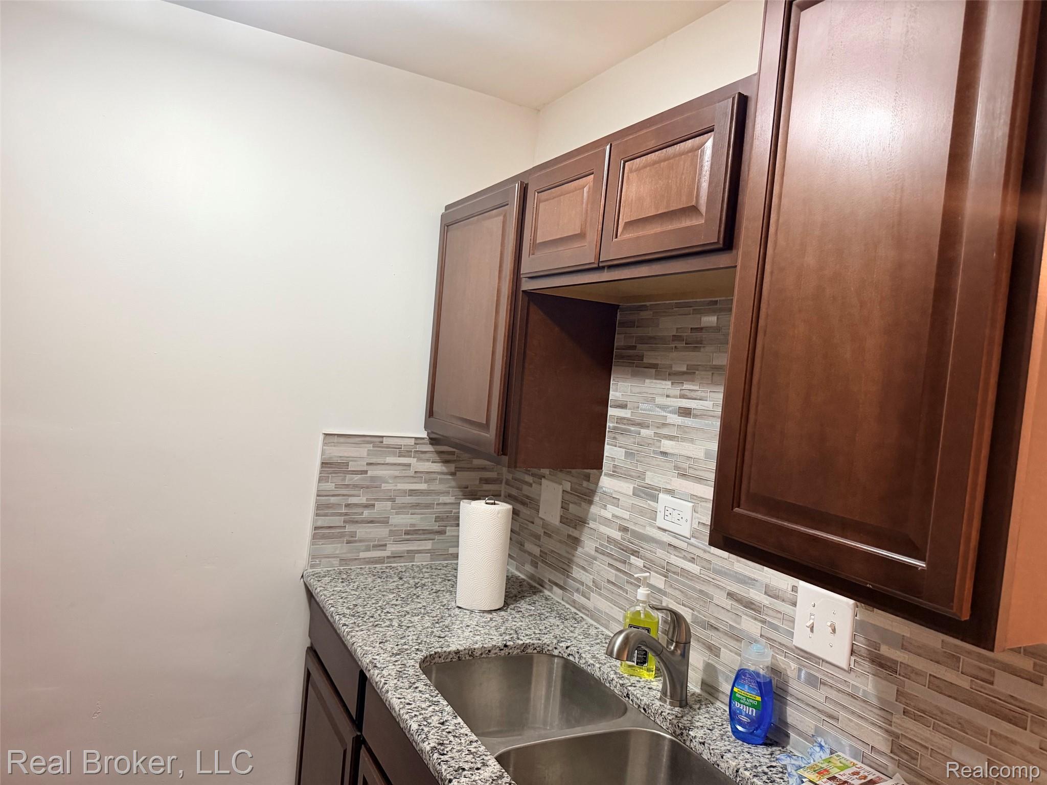 22444 Kelly Road, Unit 11 Eastpointe, MI 48021 - Photo 7 of 18 Dark wood-finish cabinetry, speckled countertop, stainless steel double basin sink, and mosaic tile backsplash