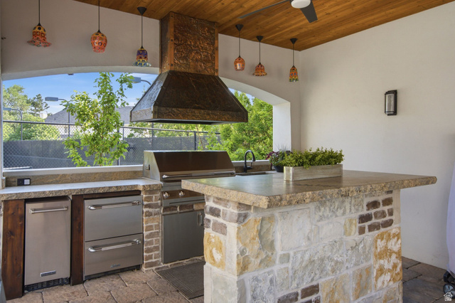 12308 Mariah Circle Draper, UT 84020 - Photo 156 of 293 Outdoor kitchen featuring a stone and brick island, built-in stainless steel grill, a decorative range hood, and a ceiling fan