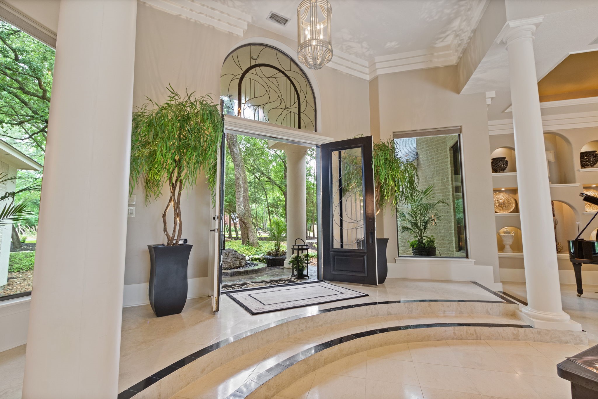 17511 Seidel Road Tomball, TX 77377 - Photo 12 of 49 This elegant foyer features high ceilings, large windows, and a grand entrance with double doors. The space is adorned with decorative columns, a stylish chandelier, and built-in shelves showcasing art pieces. Lush greenery outside enhances the inviting atmosphere.