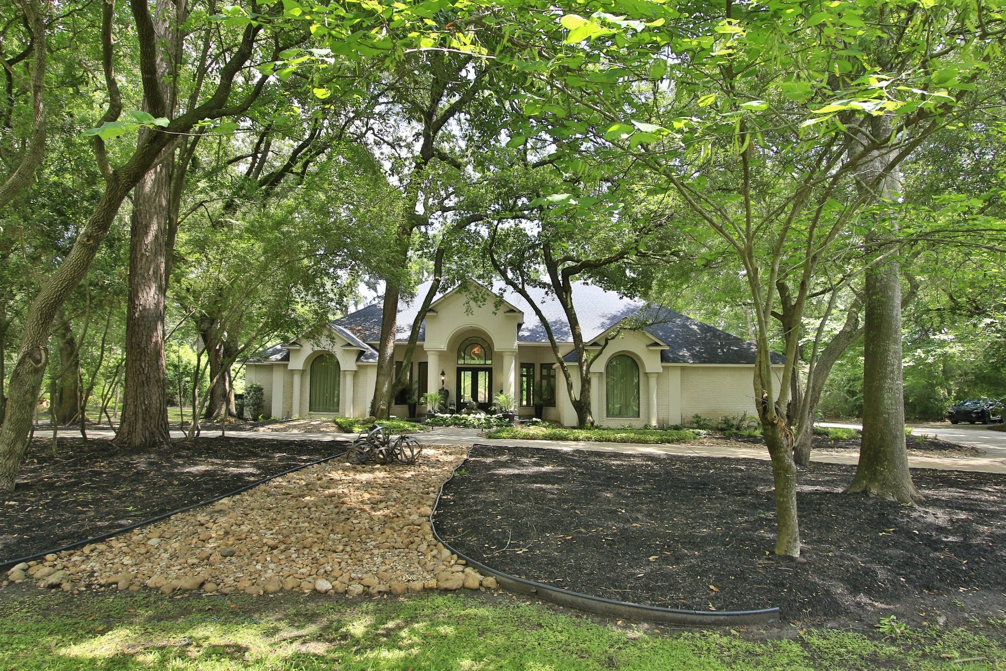 17511 Seidel Road Tomball, TX 77377 - Photo 41 of 49 This photo features a charming single-story home nestled among mature trees, offering a sense of privacy and tranquility. The house has a welcoming front entrance with large windows and a well-maintained landscape, including a neat rock pathway and lush greenery. Perfect for those seeking a serene, nature-surrounded living space.