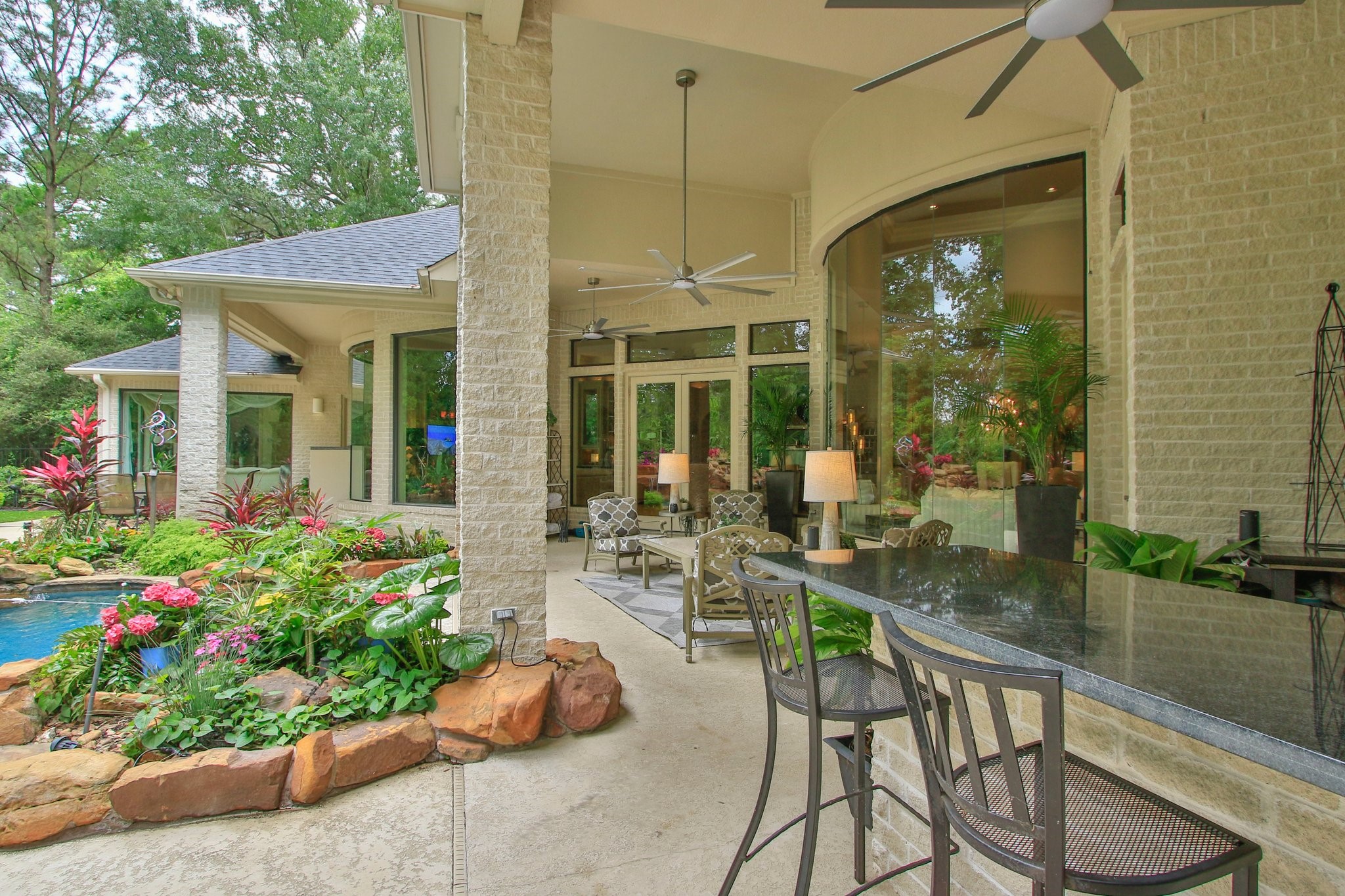 17511 Seidel Road Tomball, TX 77377 - Photo 8 of 49 This photo showcases a beautiful outdoor living space with a covered patio featuring ceiling fans and ample space for your outdoor furniture. There's a stone bar for entertaining, and large windows provide a view into the lush garden and pool area, surrounded by vibrant landscaping. Ideal for relaxation and gatherings.