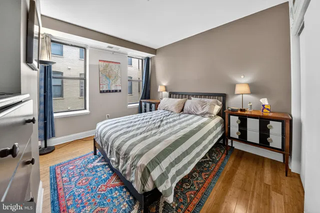 a bedroom with a bed and a lamp on dresser with wooden floor