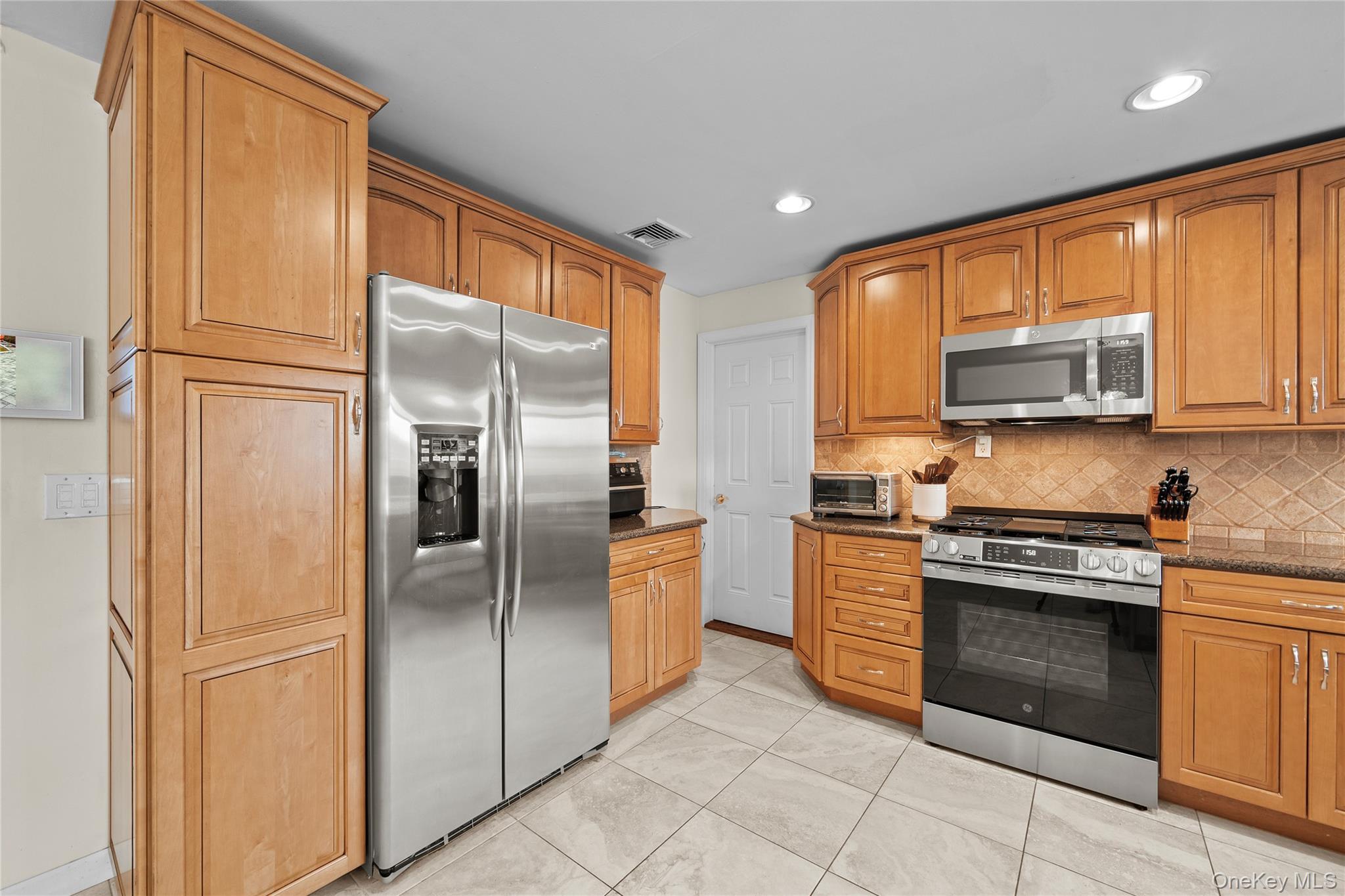 625 Edgewater Road Oakdale, NY 11769 - Photo 15 of 49 Kitchen with stainless steel appliances, backsplash, dark stone counters, light tile patterned floors, and brown cabinetry