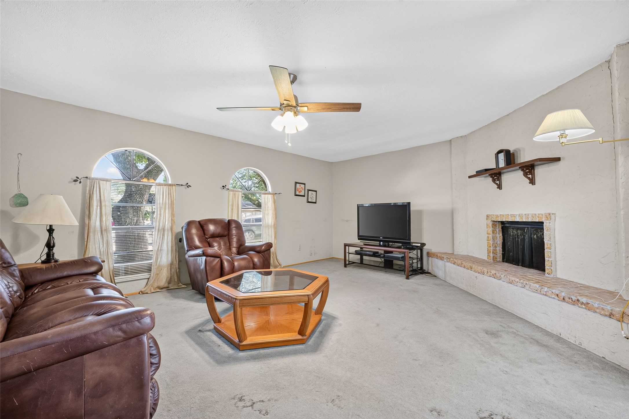 22926 Millgate Drive Spring, TX 77373 - Photo 7 of 34 This inviting living room features a cozy fireplace, ample seating with leather sofas, and a ceiling fan for comfort. Natural light pours in through arched windows, complementing the neutral walls. A modern TV setup and a unique hexagonal coffee table complete the space, perfect for relaxation and entertainment.