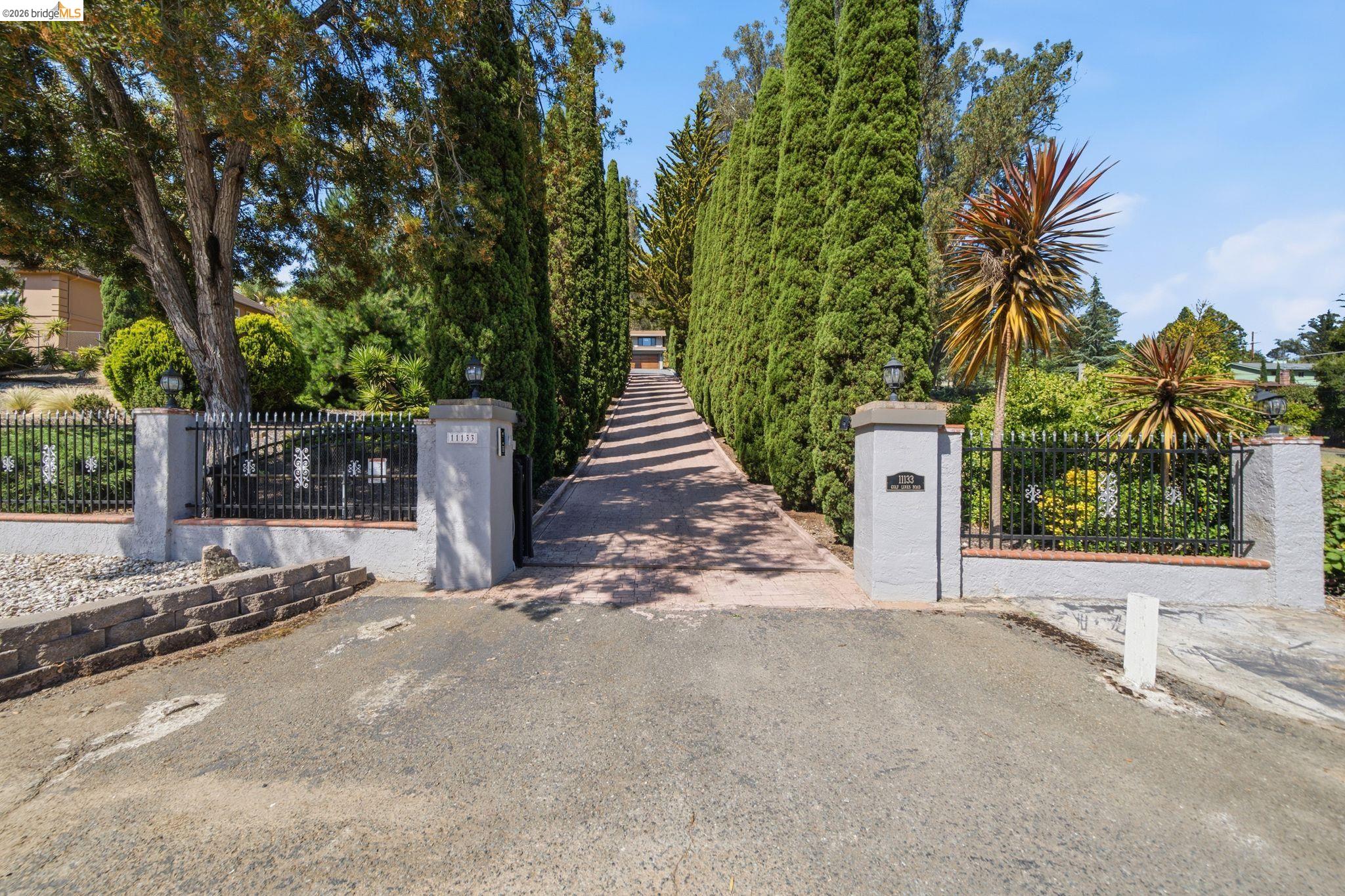 11133 Golf Links Road Oakland, CA 94605 - Photo 2 of 57 Gated entry with stone pillars, wrought iron fencing, and a paved driveway bordered by mature evergreen trees
