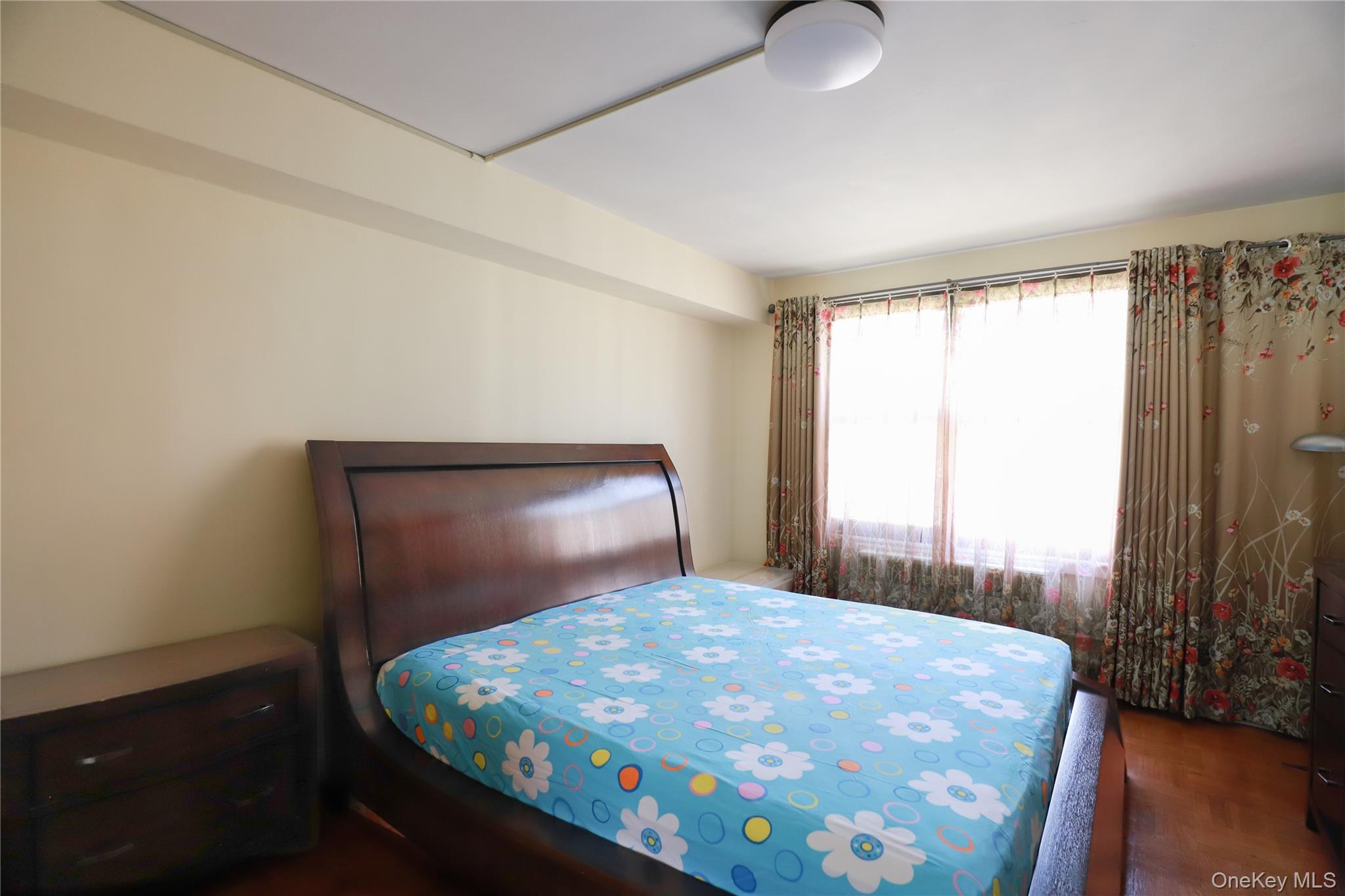 98-25 64th Road, Unit 6F Queens, NY 11374 - Photo 8 of 12 Bright interior bedroom featuring a large window with sheers and patterned drapes, wood-finish flooring, and overhead lighting