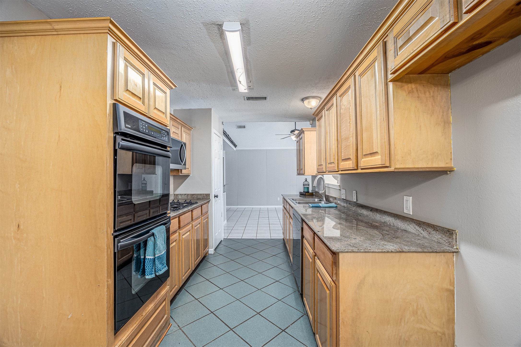 1603 East Baker Road Baytown, TX 77521 - Photo 11 of 26 This photo showcases a galley kitchen with natural wood cabinetry, granite countertops, and modern appliances. The space is well-lit with overhead lighting and features a tile floor, leading into an open area with tile flooring.