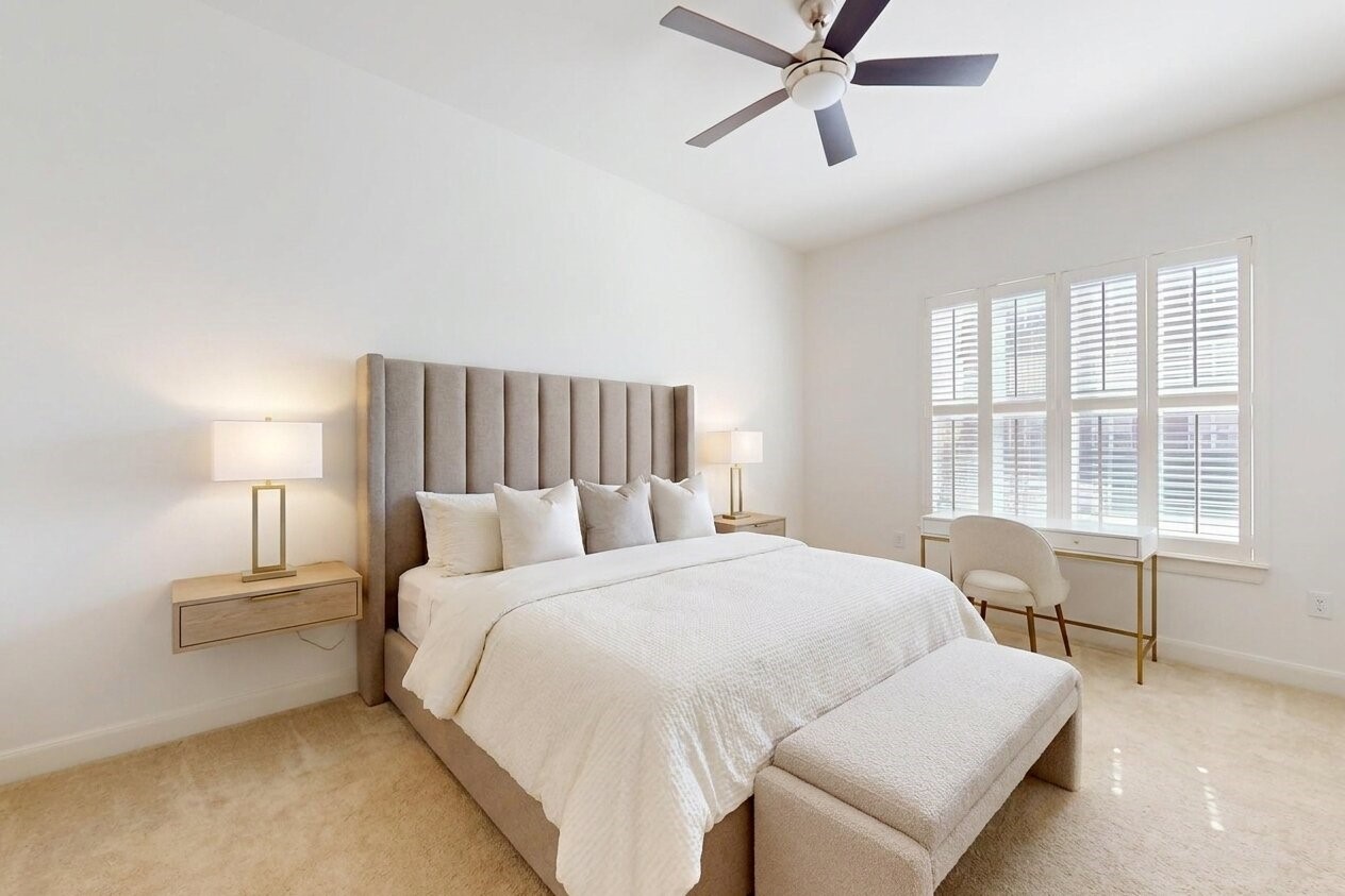1010 Rosine Street, Unit 29 Houston, TX 77019 - Photo 10 of 16 This bright and airy bedroom features a plush bed with a tall upholstered headboard, cozy carpeting, and a modern ceiling fan. Large windows with shutters allow natural light, while dual side tables and a sleek desk provide functionality. (Virtual Staging)