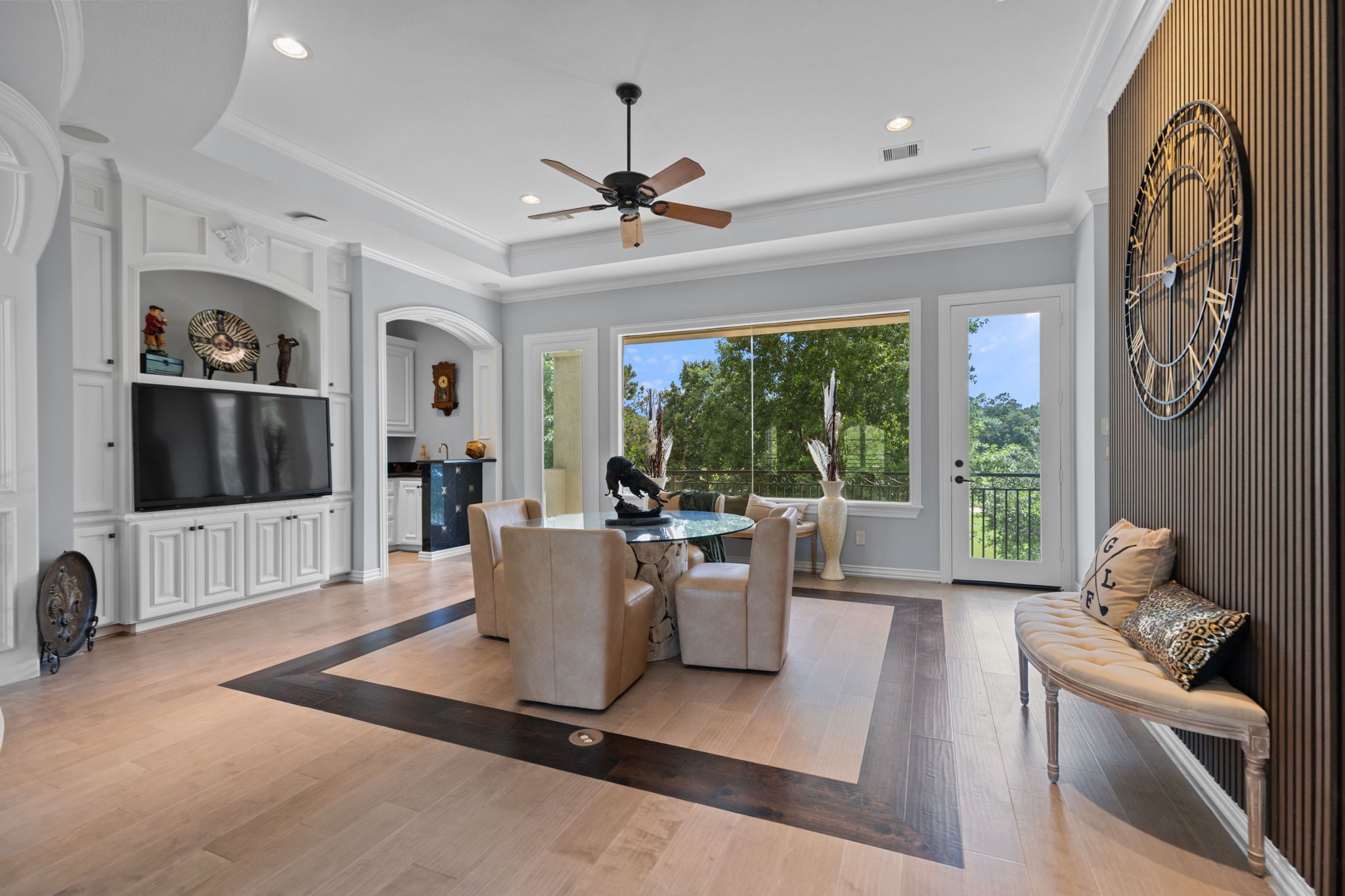 39 Maymont Way Spring, TX 77382 - Photo 26 of 50 This spacious living area features elegant hardwood floors, a large TV with built-in cabinetry, and a modern ceiling fan. The room has ample natural light from expansive windows and a door leading to a scenic view. A stylish dining area with plush chairs and a glass table sits near a wall with a decorative clock.