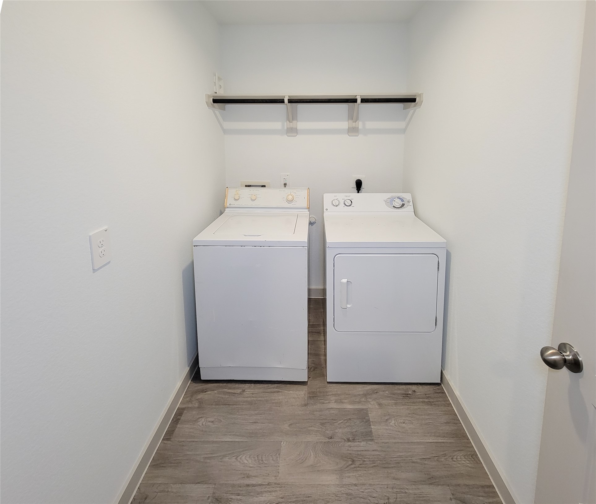 7722 Caldwell Crk Way Rosharon, TX 77583 - Photo 29 of 40 This photo shows a compact laundry room with a washer and dryer side by side. It features light-colored walls, wood-style flooring, and a shelf above for storage.