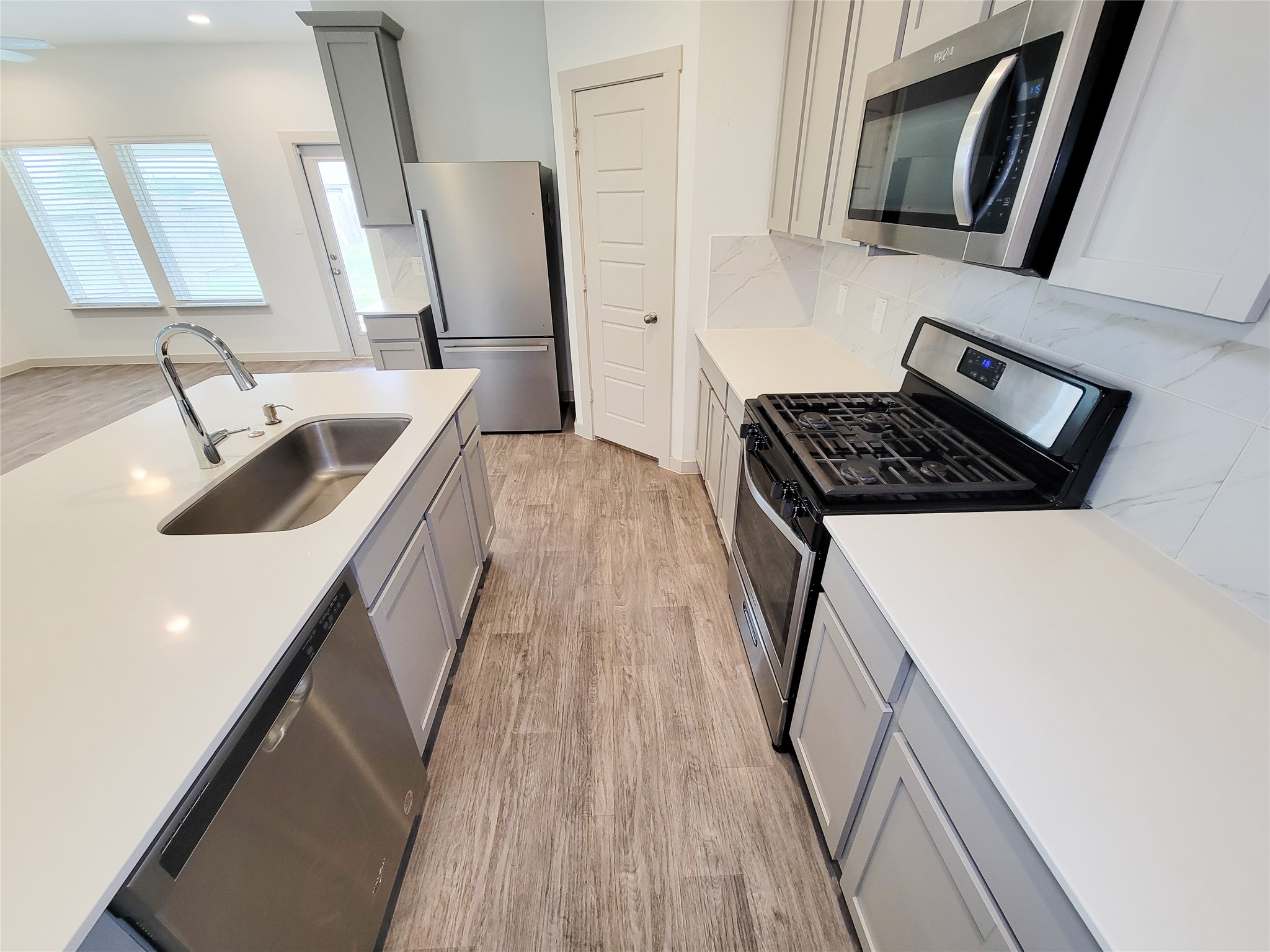 7722 Caldwell Crk Way Rosharon, TX 77583 - Photo 10 of 40 Modern kitchen with stainless steel appliances, white countertops, and light-colored cabinetry. Features a central island with a sink and dishwasher, wood-look flooring, and ample natural light from nearby windows.