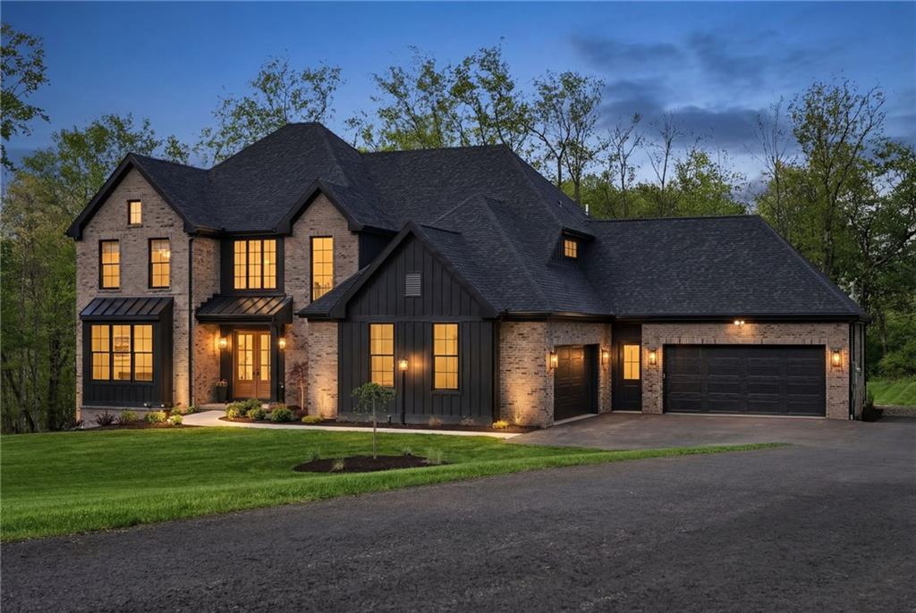 225 Stoup Road at dusk — warm light spills from every window, highlighting the brick and board-and-batten exterior, standing seam metal awnings, and four-car garage against a deep blue evening sky. A home that makes just as strong an impression after dark.