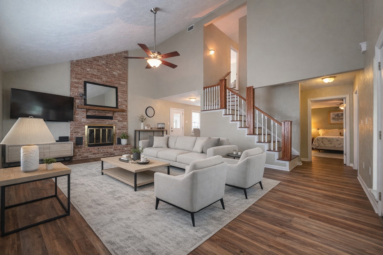 Spacious living room with vaulted ceilings, featuring a cozy brick fireplace and stylish wood flooring. An elegant staircase leads to the upper level, with natural light enhancing the open layout.