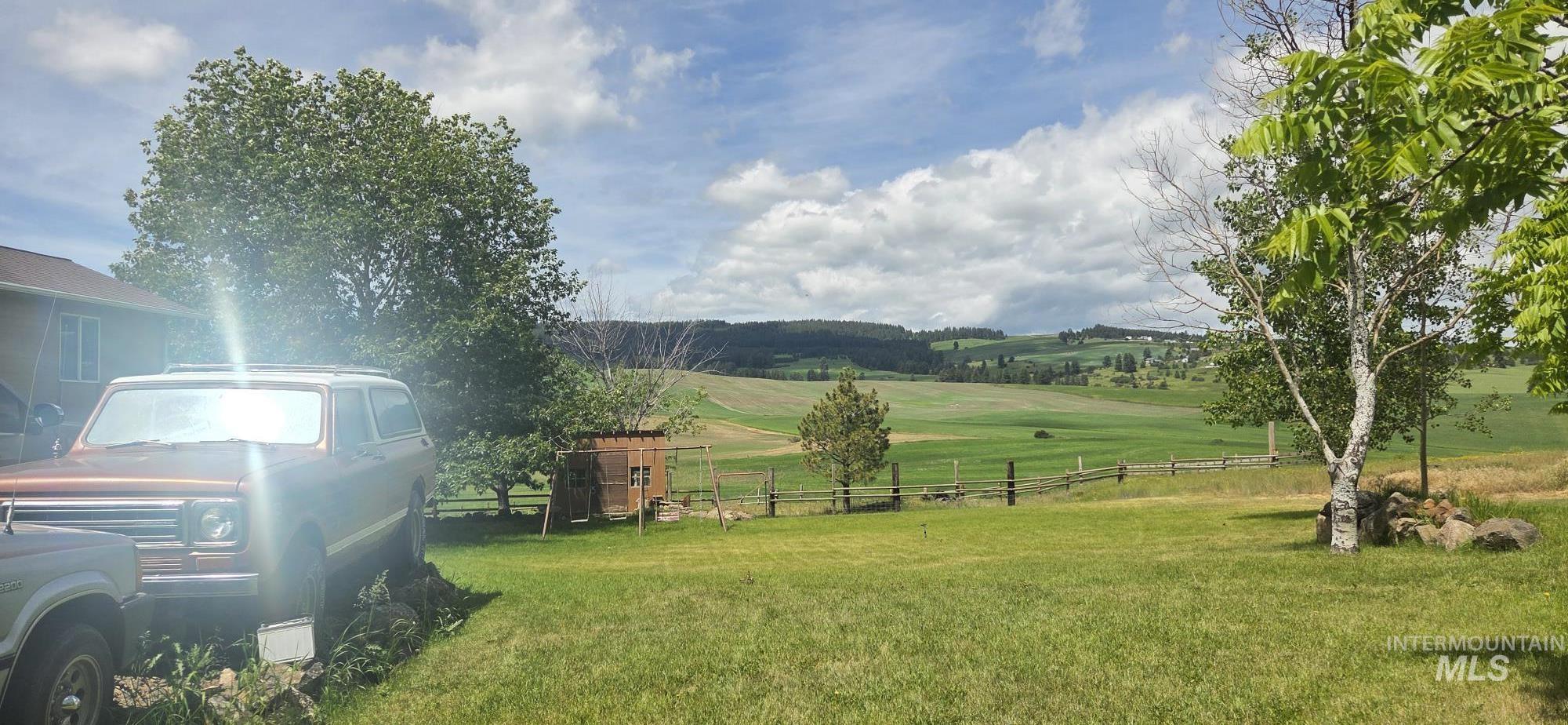394 Substation Road Cottonwood, ID 83522 - Photo 12 of 37 Expansive green landscape with distant rolling hills, featuring a rustic wood fence, mature trees, and a natural stone retaining wall