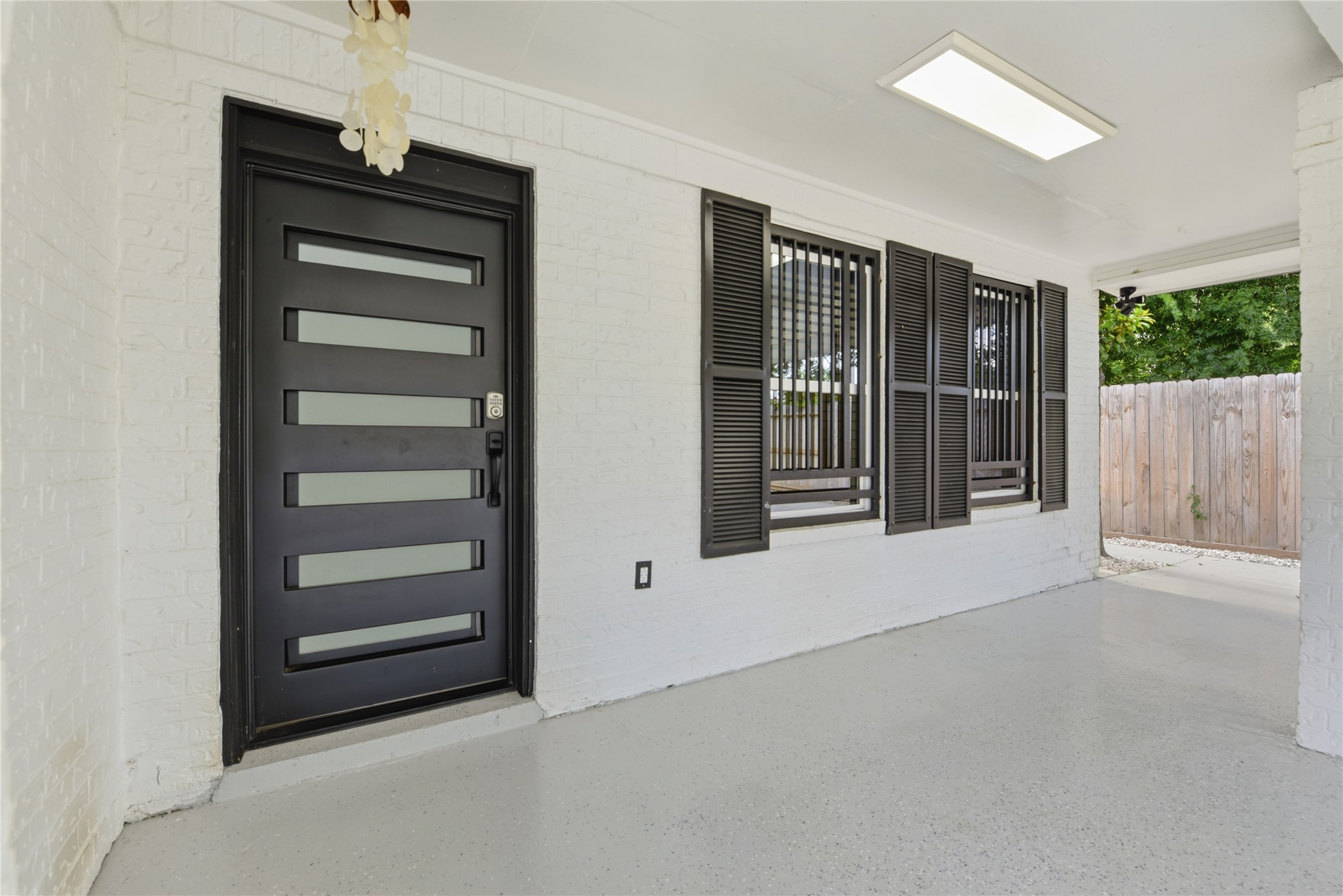 9403 Spaulding Street Houston, TX 77016 - Photo 3 of 50 This photo showcases a modern, covered entryway with a sleek black door featuring horizontal glass panels. The area has white brick walls, black shutters on the windows, and a light gray floor. There's a wooden fence visible, adding privacy.