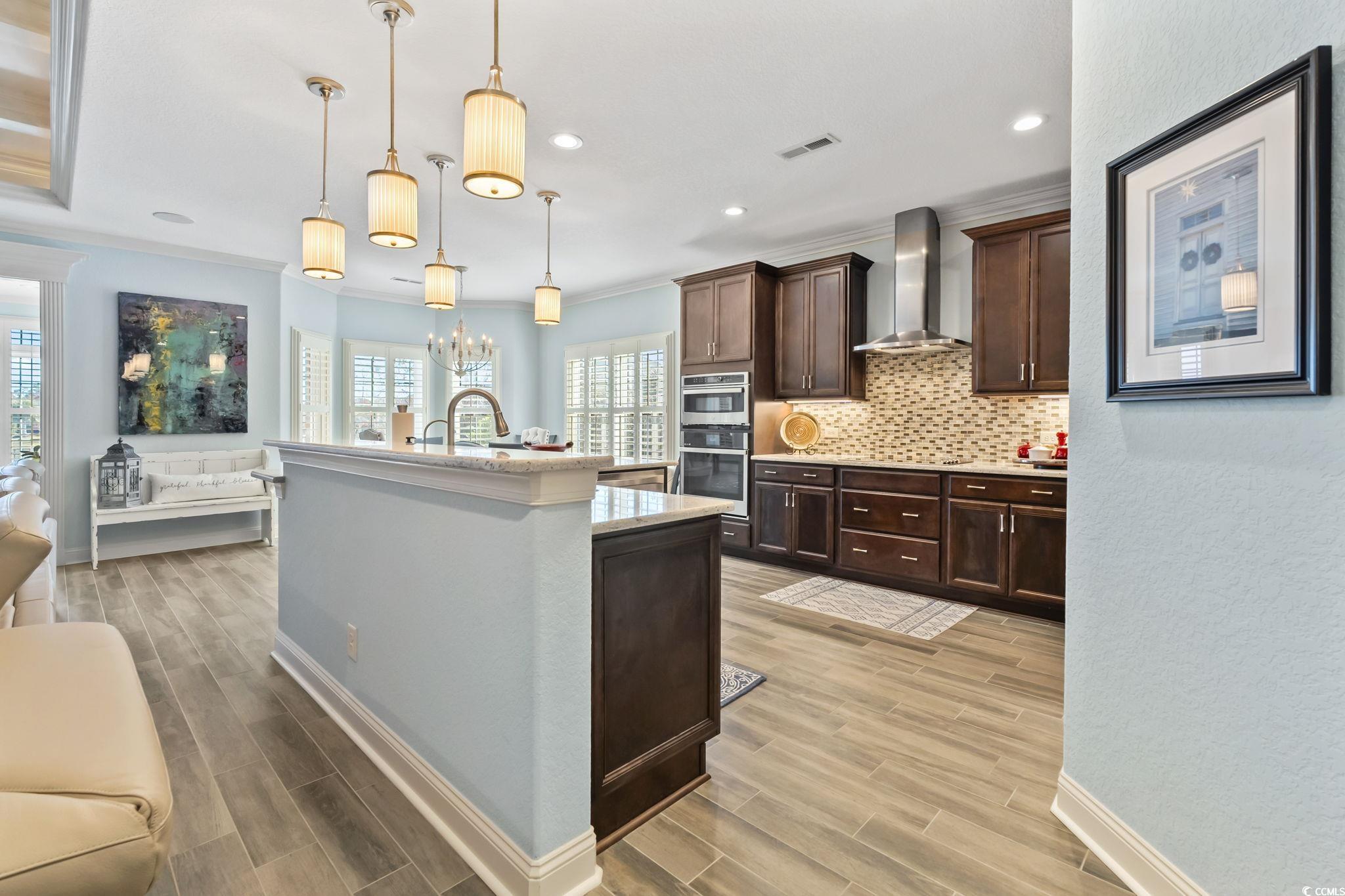 750 Villarosa Drive Myrtle Beach, SC 29572 - Photo 11 of 40 Kitchen featuring hanging light fixtures, light stone counters, dark brown cabinetry, wall chimney exhaust hood, and ornamental molding