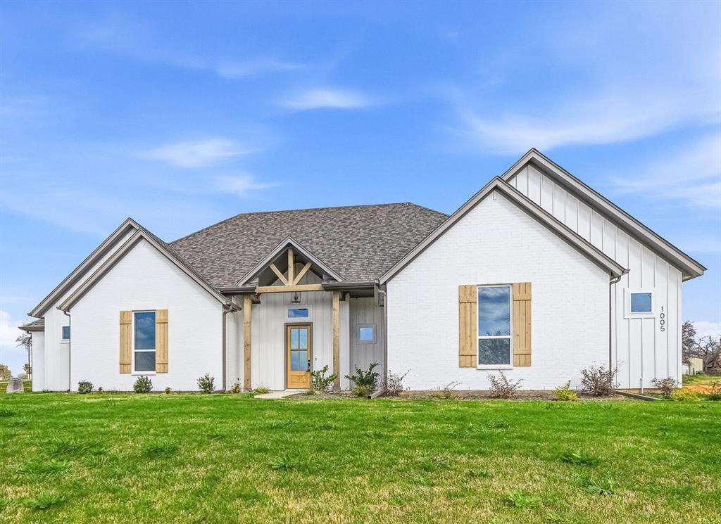 1005 Ronnie Court Springtown, TX 76082 - Photo 3 of 27 Modern farmhouse-style exterior featuring white brick and vertical siding, a covered entry with exposed wood beams, a prominent front door with glass inserts, and decorative wooden shutters flanking multiple windows