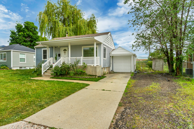 65 West 700 North Bountiful, UT 84010 - Photo 4 of 29 Light blue siding exterior featuring a covered front porch with white railings, a concrete driveway, a single-car garage, and mature trees