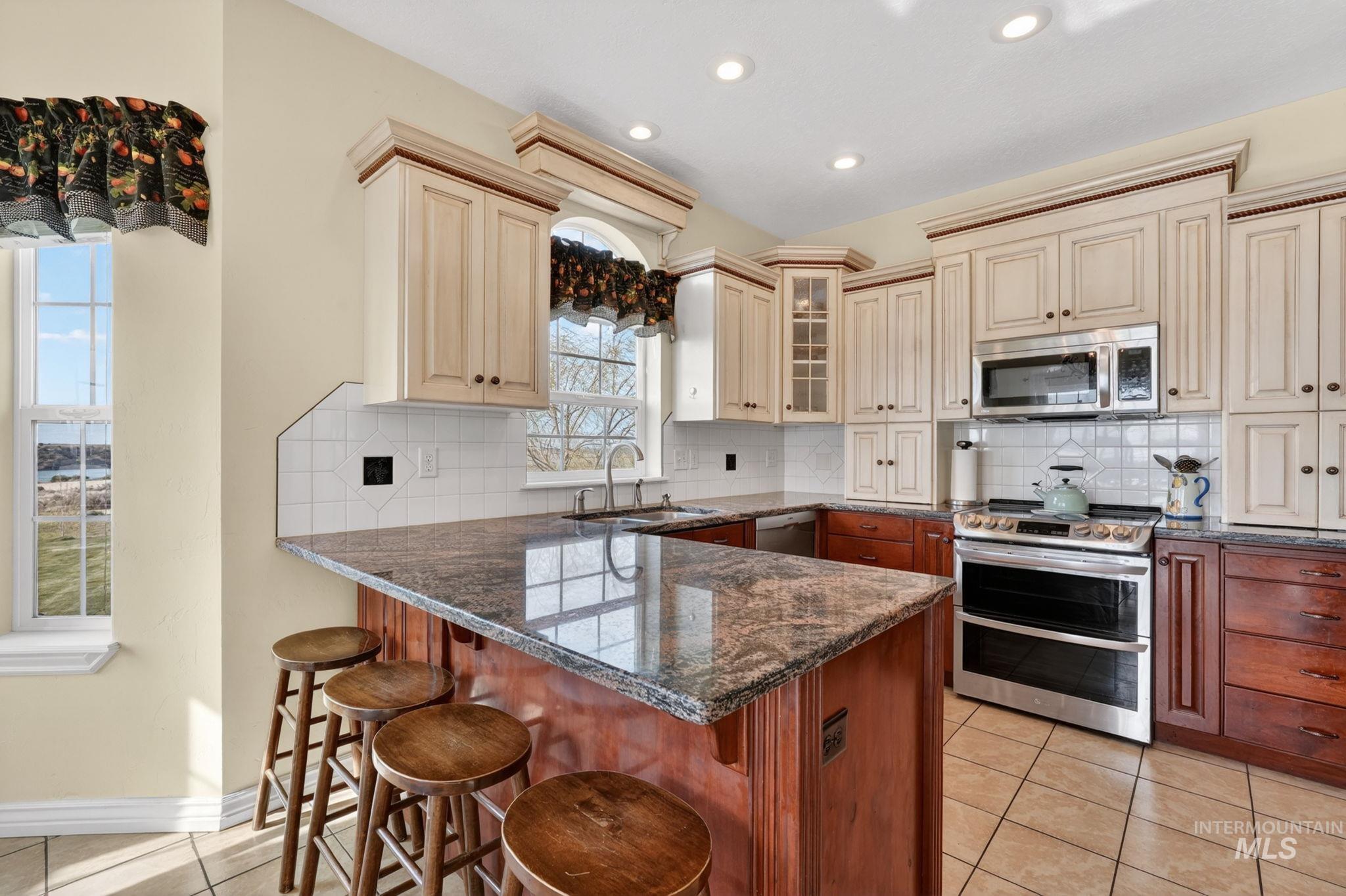 3352 Neeley Road American Falls, ID 83211 - Photo 10 of 43 Kitchen featuring a large central island with seating, granite countertops, tile backsplash, stainless steel appliances, and a mix of light-toned upper cabinetry and wood-finish lower cabinetry