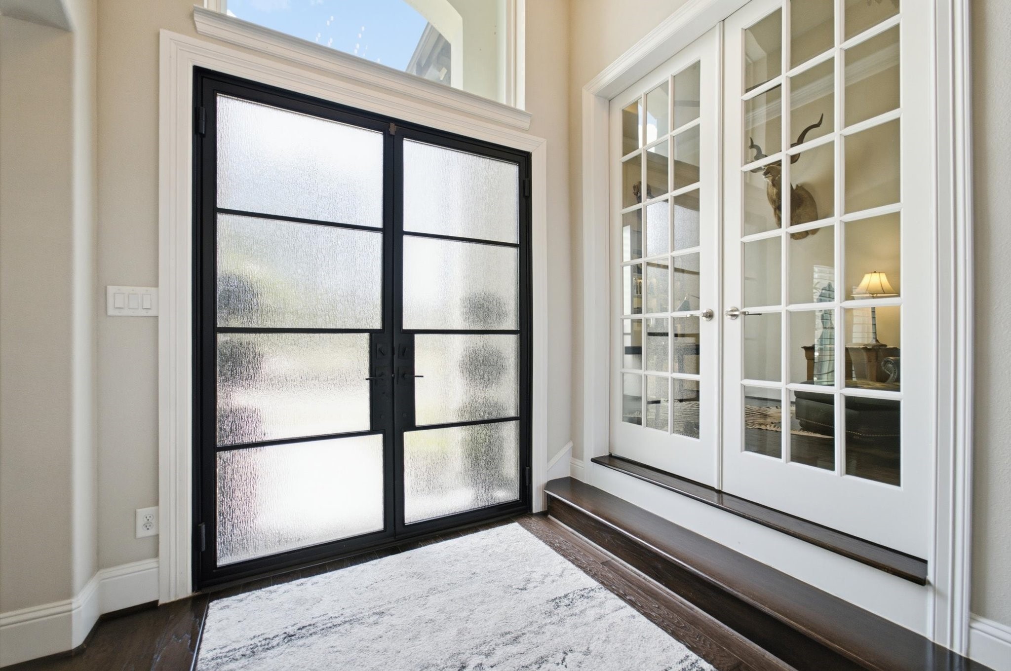 23722 Thortons Park Lane Spring, TX 77389 - Photo 5 of 48 This entryway features elegant frosted rain glass double doors with black frames, leading to a bright interior. Adjacent are French doors with glass panes, providing a view into a cozy, well-lit room. The space is accentuated with a light area rug and dark hardwood flooring.