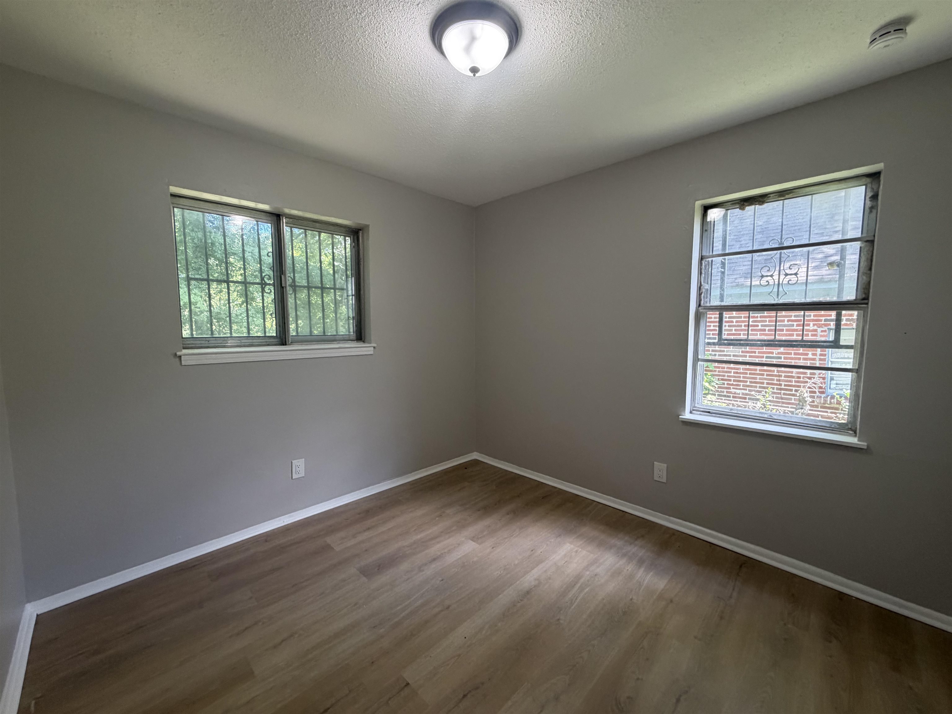 2402 Shasta Avenue Memphis, TN 38108 - Photo 5 of 7 Room featuring wood-finish flooring, two windows with security bars, painted gray walls, white baseboards, and a flush-mount ceiling light