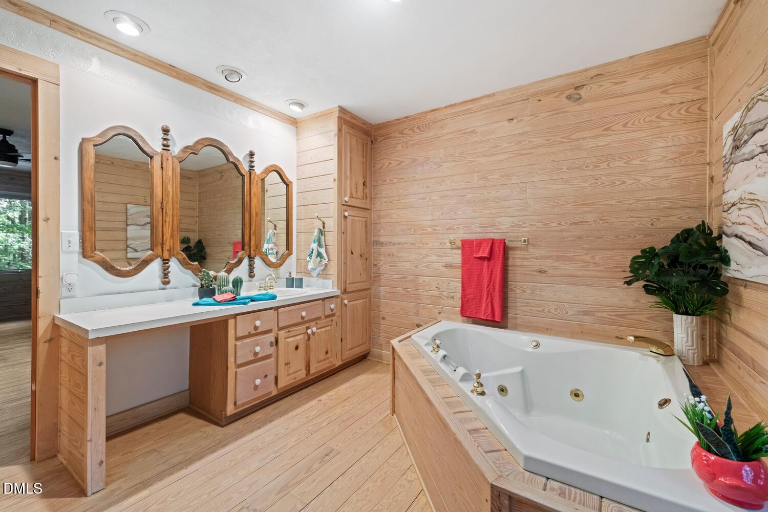 7391 Howerton Road Elon, NC 27244 - Photo 41 of 53 Wood-finish flooring with wood-paneled walls, built-in vanity with integrated sink, three ornate wood-framed mirrors, and a white jetted tub with gold-tone fixtures