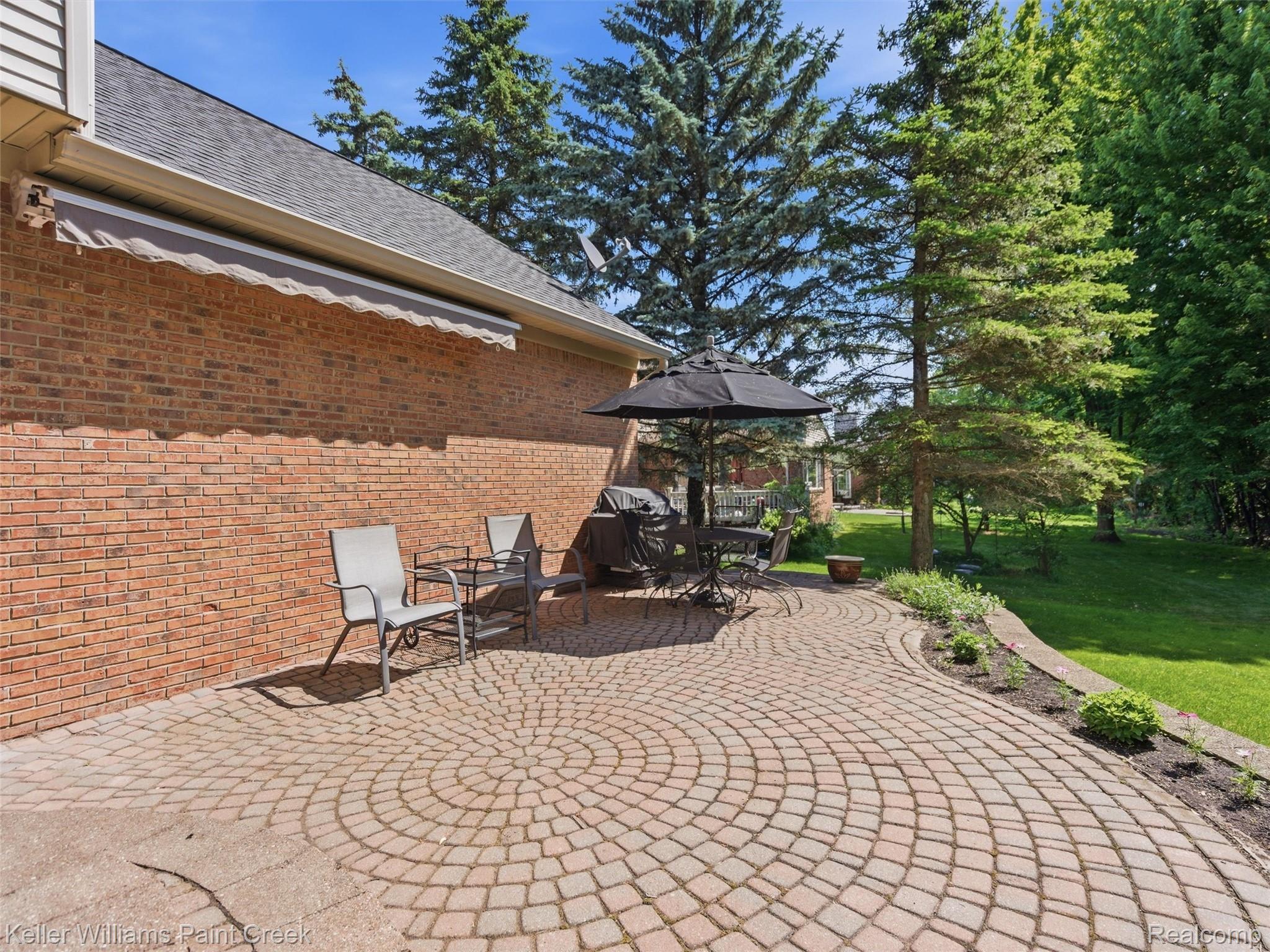 2842 Manorwood Road Troy, MI 48085 - Photo 69 of 72 Expansive paver patio featuring a circular pattern, bordered by a raised garden bed with plantings, and a brick exterior wall with an attached retractable awning